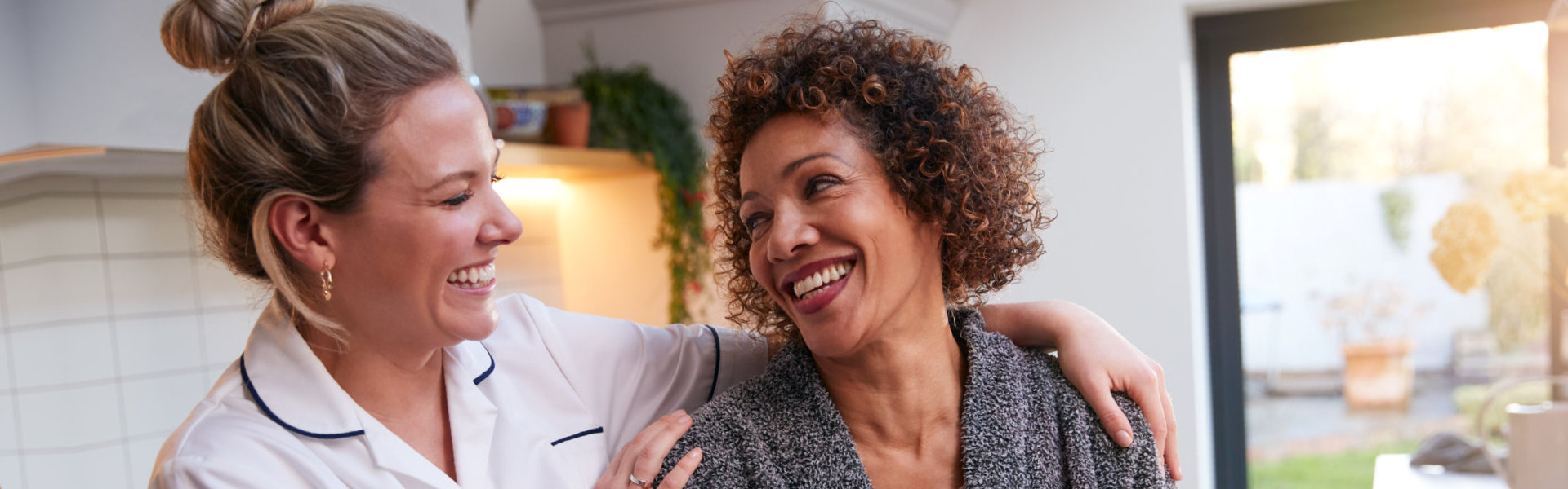 Mature Woman In Dressing Gown Talking With Female Nurse In Kitchen At Home
